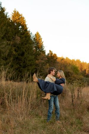A couple embraces in a romantic dip pose against a backdrop of autumn trees and tall grass at sunset.