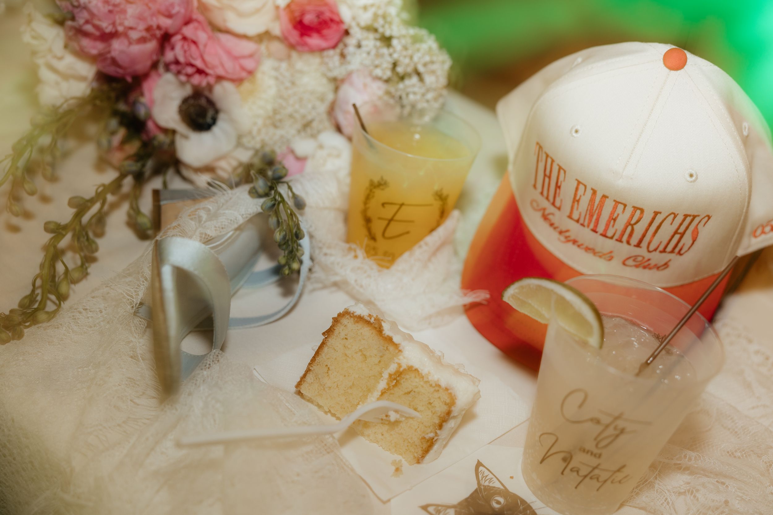 Wedding table with floral bouquet, cake slice, personalized cups, and The Emericks hat on lace tablecloth.