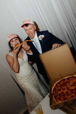 Bride and groom in matching pizza restaurant caps joyfully eating pepperoni pizza slices at their wedding reception.