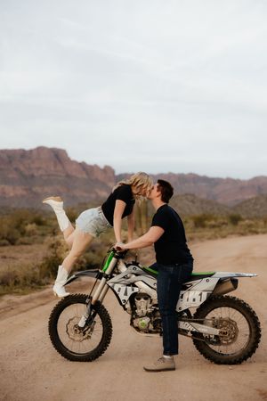 A couple embraces romantically on a dirt bike in the desert with mountains in the background.