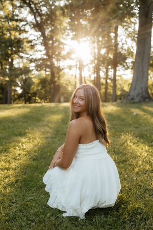 Young woman in white strapless dress sitting on grass in sunlit park, smiling over shoulder at golden hour.