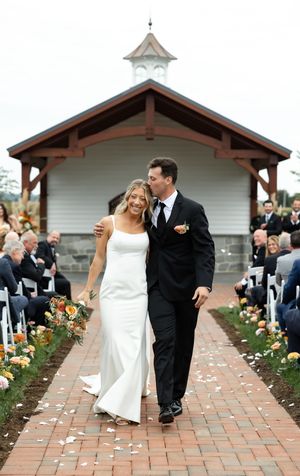 Joyful couple exits outdoor chapel ceremony on brick walkway lined with flowers.