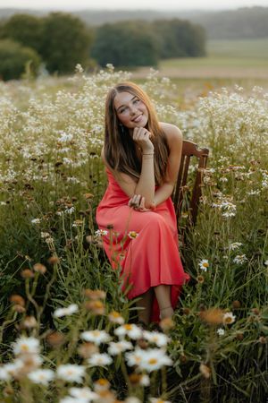 A person in a coral dress sits among blooming white daisies in a summer meadow during golden hour.