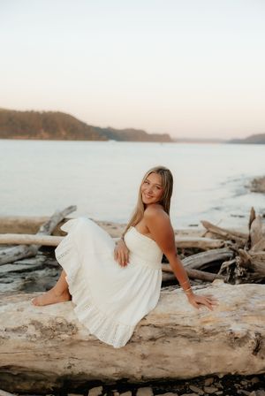 Young woman in white dress sitting on driftwood at beach during golden hour sunset.