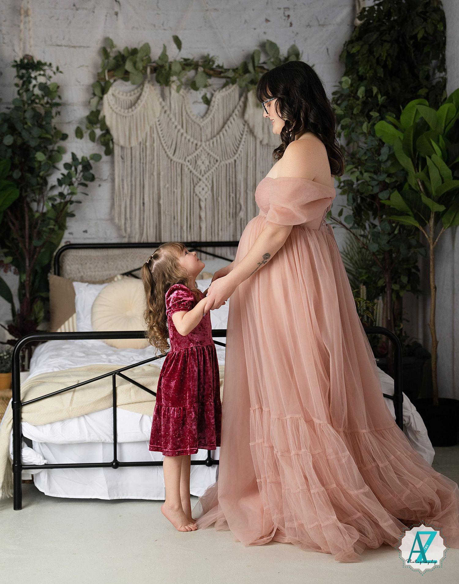 A woman in a flowing pink gown stands beside a bed with macrame wall decor and greenery.