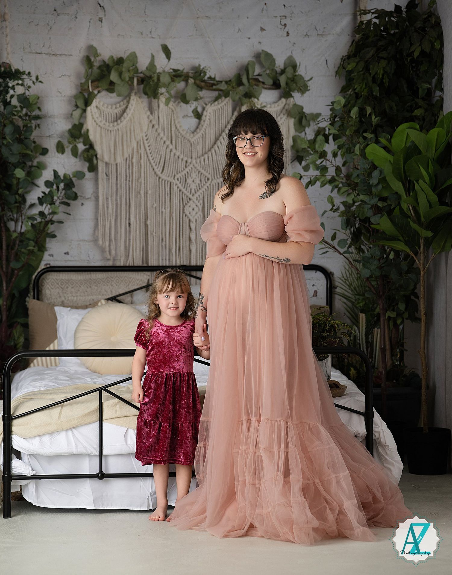 A maternity portrait features a peach tulle gown and burgundy dress against a macrame backdrop with greenery.
