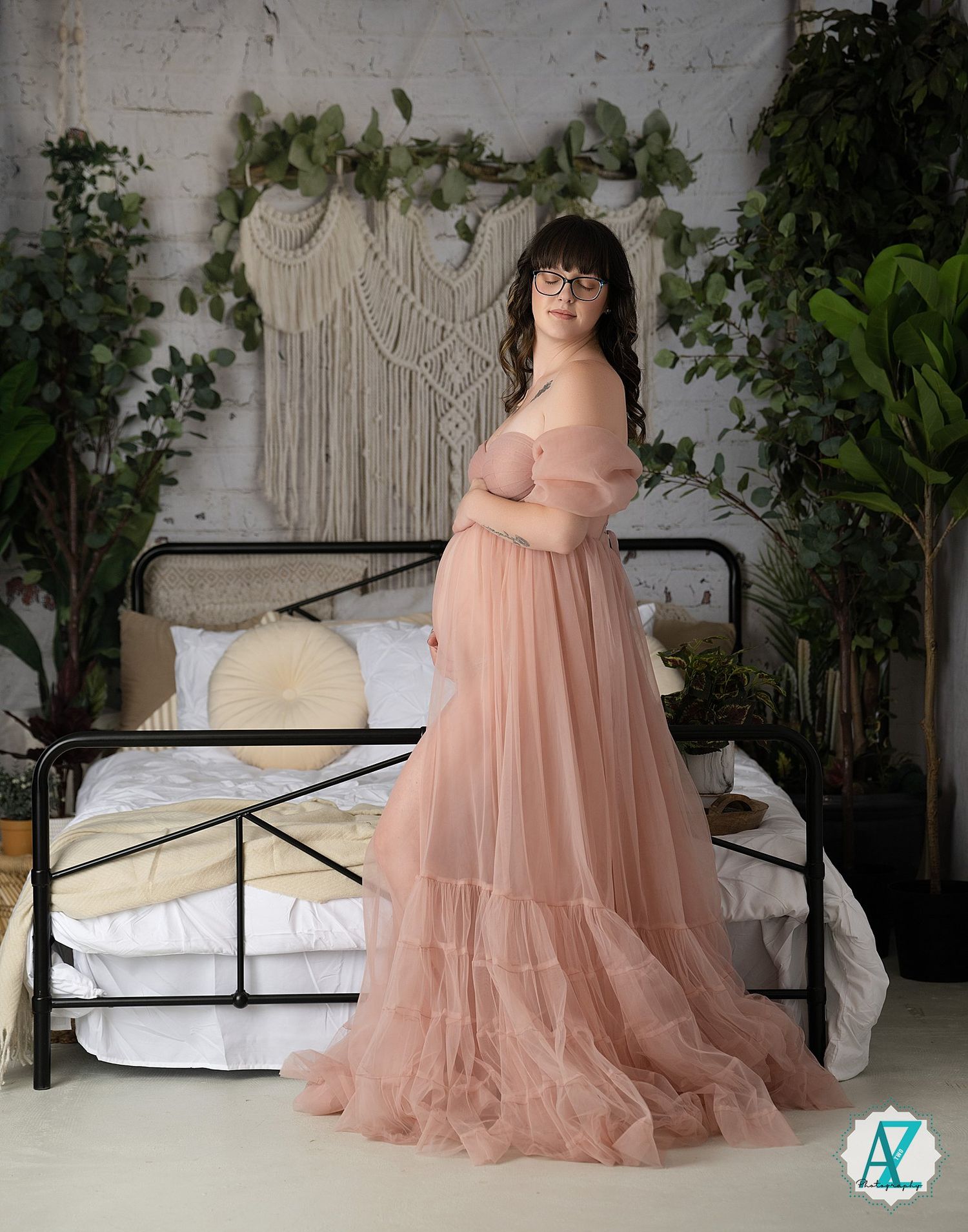 A romantic maternity photoshoot features a flowing pink tulle gown against a white backdrop with green foliage accents.