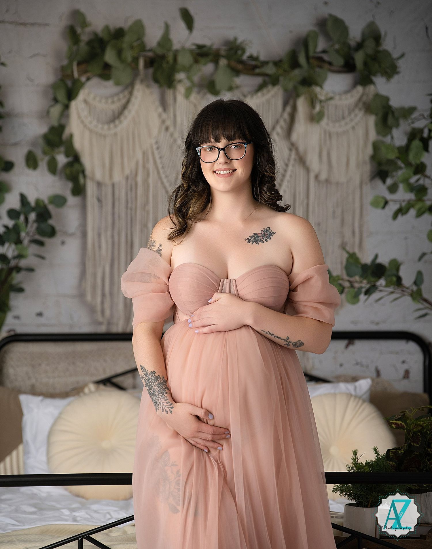 A maternity portrait featuring a soft pink off-shoulder gown against a macrame wall hanging with ivy.