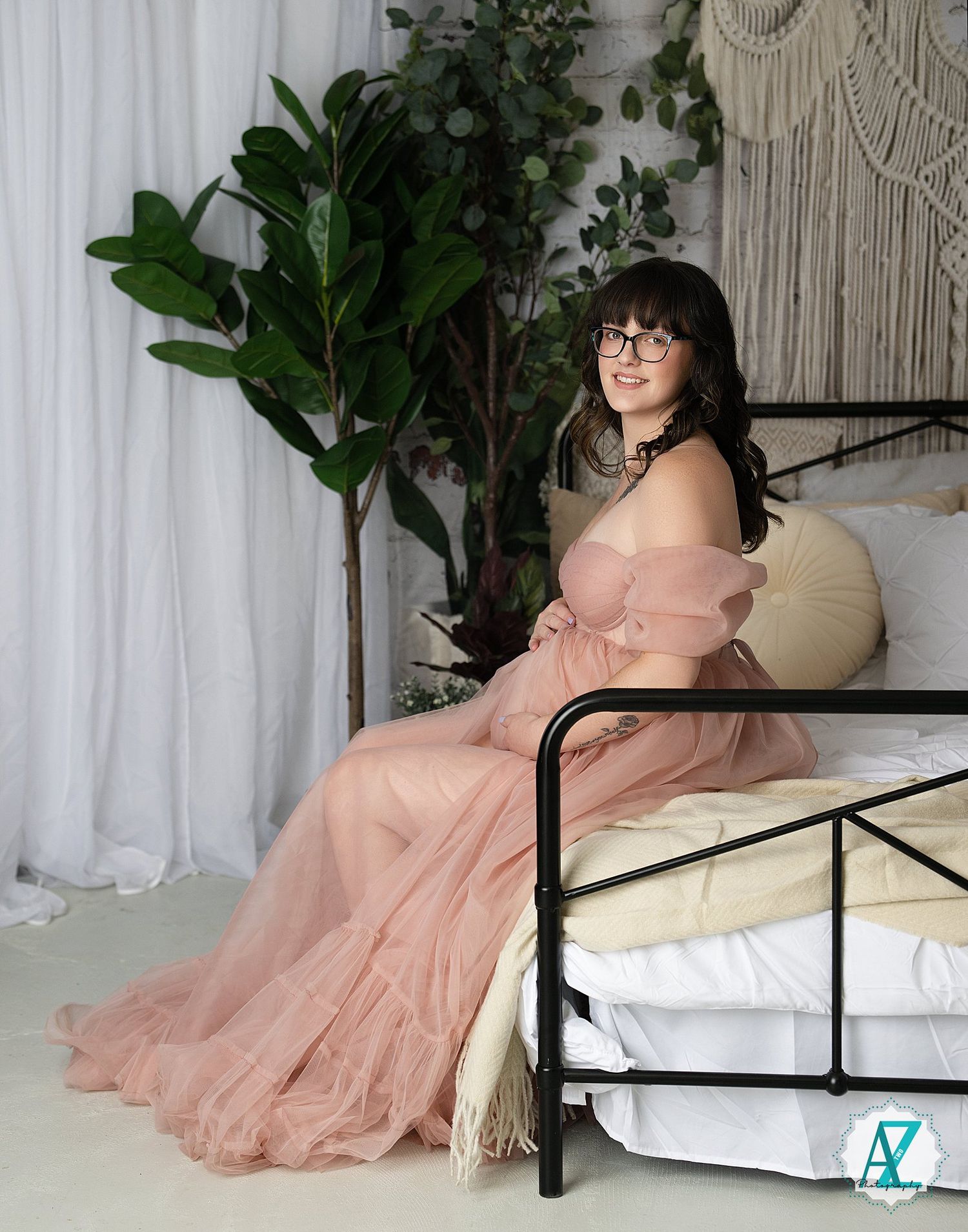 A person in a flowing peach-colored gown sits on a bed with white linens, surrounded by plants and macrame decor.