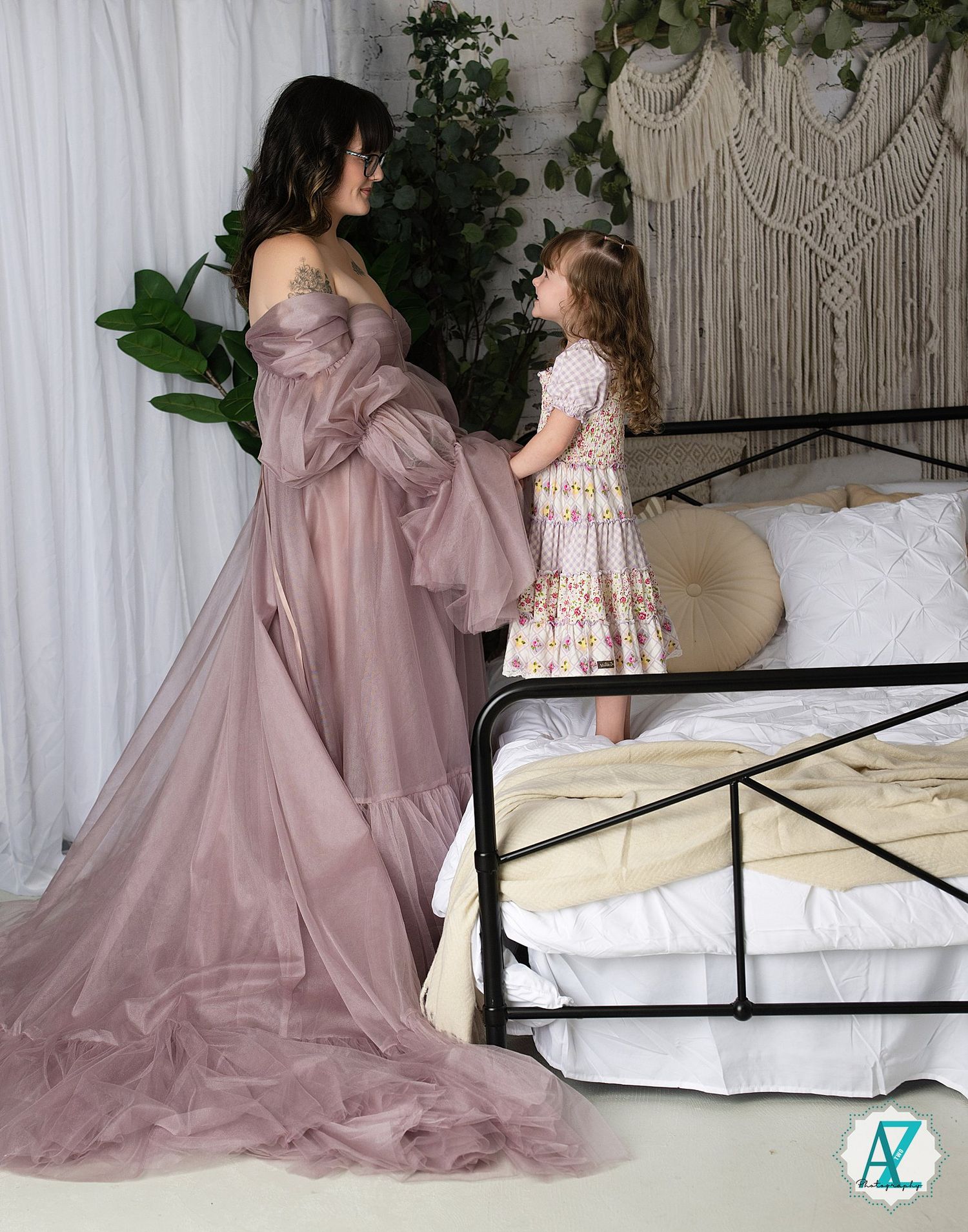 Touching maternity moment in a dreamy bedroom setting with flowing mauve dress and decorative macrame backdrop.