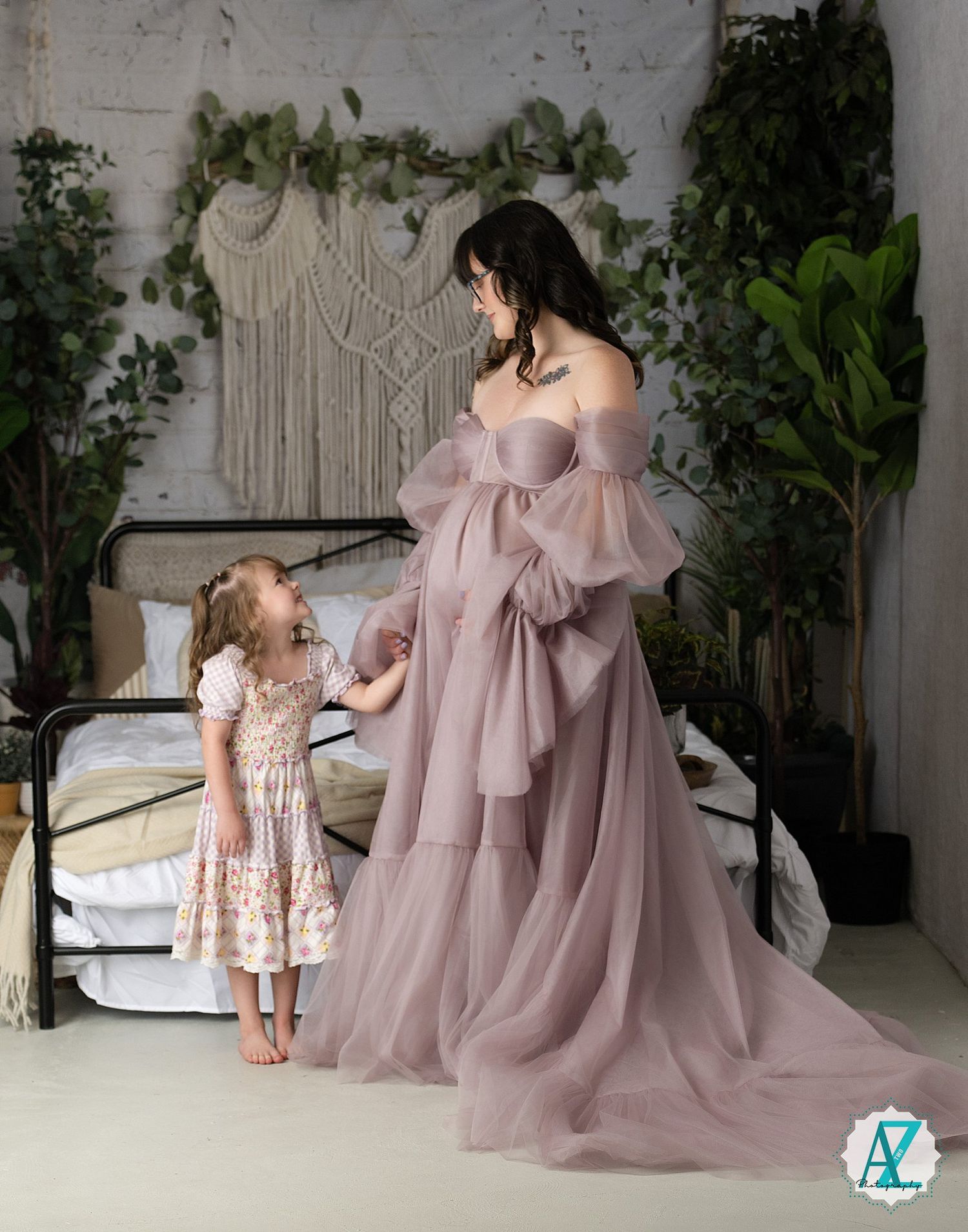 A parent and child in matching pink dresses share a tender moment in a dreamy indoor studio setting with greenery.