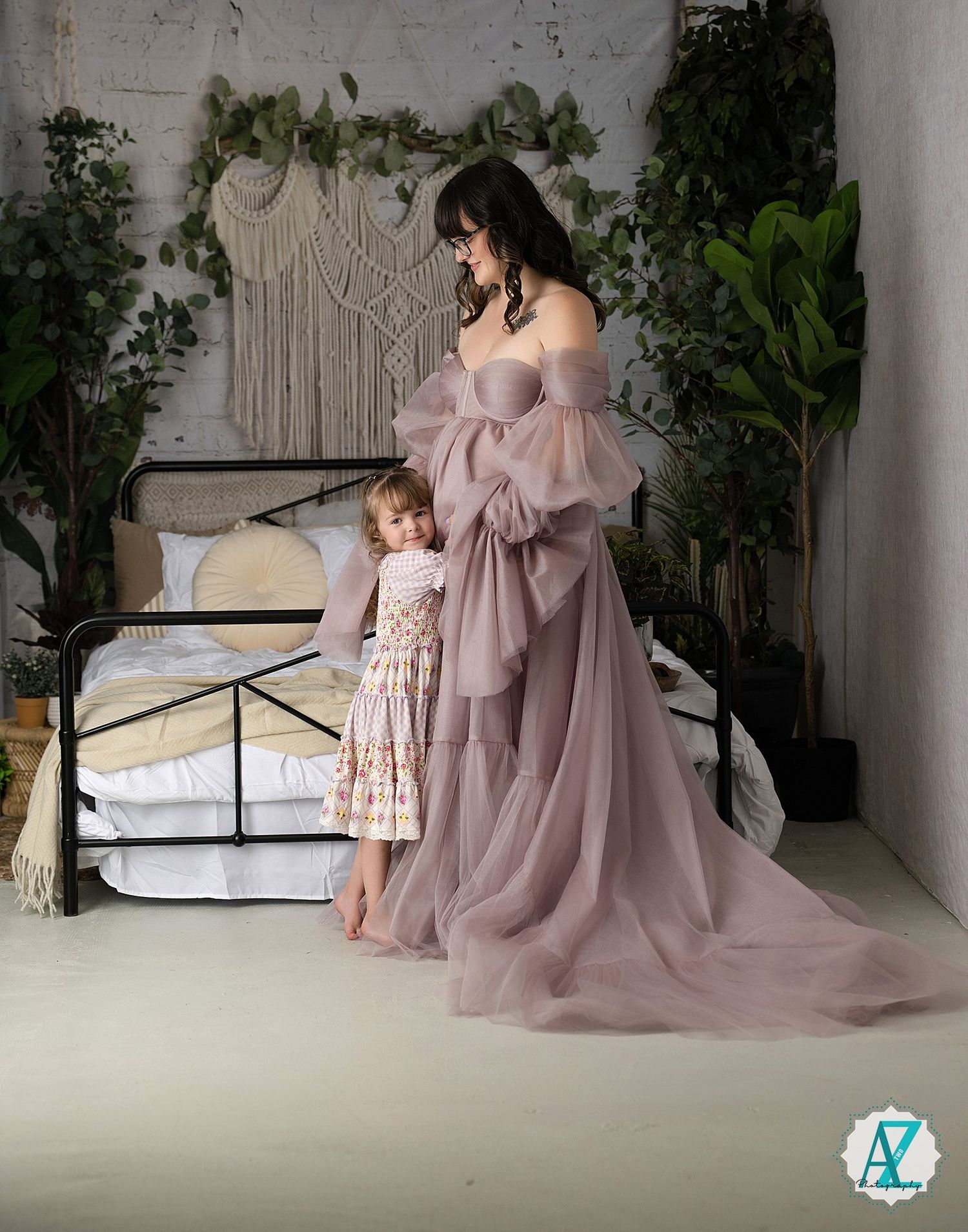 A touching maternity portrait showing matching dusty rose dresses in an intimate indoor setting with macrame decor.