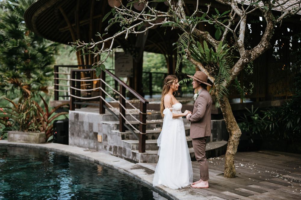 Wedding couple standing by resort pool with natural stone steps and tropical landscaping.