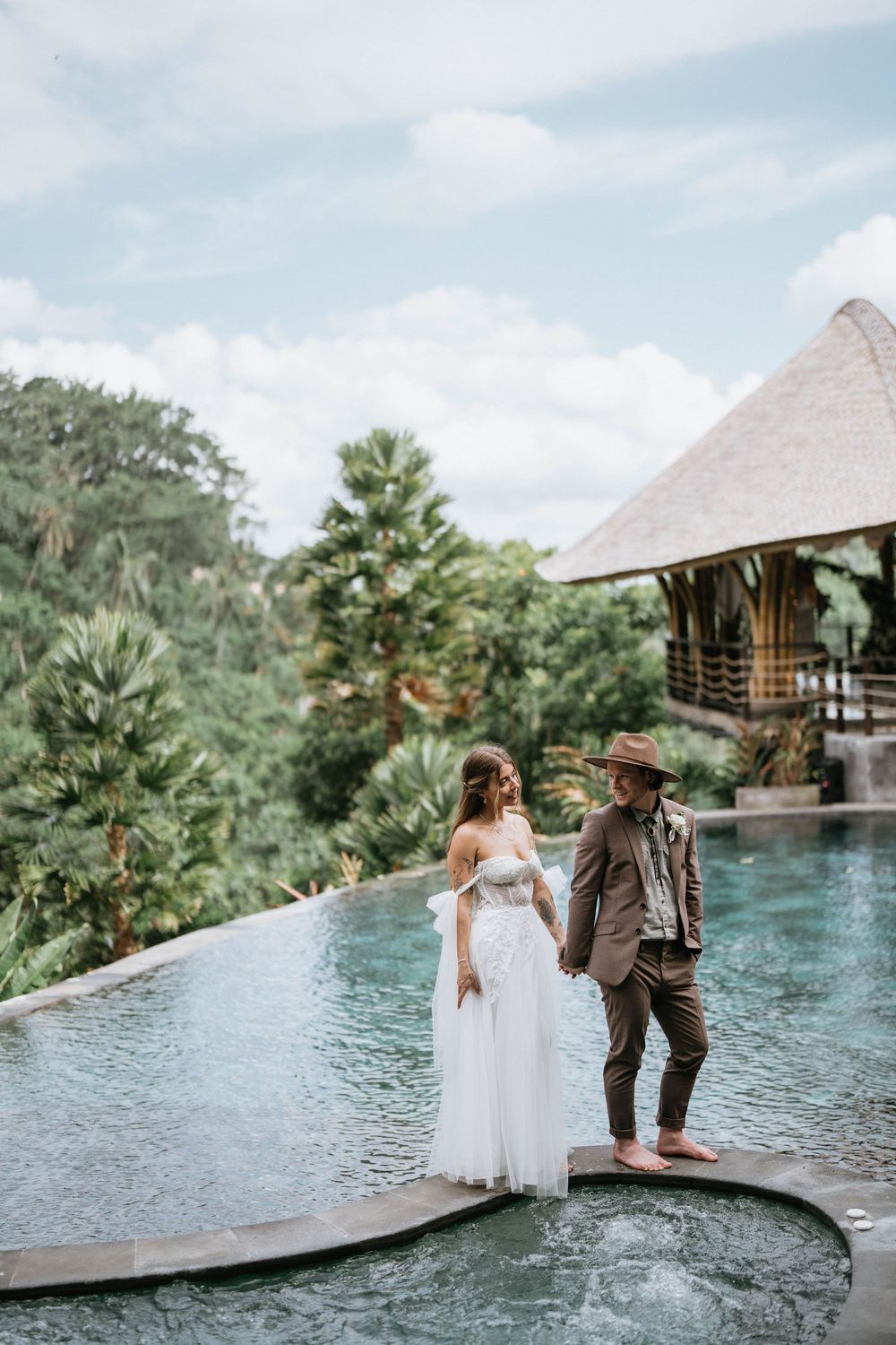 A romantic couple poses by a tropical infinity pool surrounded by lush greenery and palm trees.