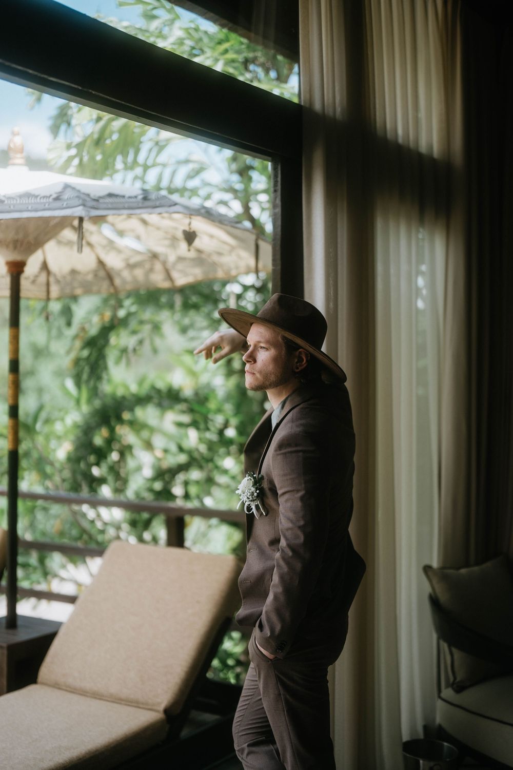 A figure in brown suit stands by large windows overlooking tropical scenery.