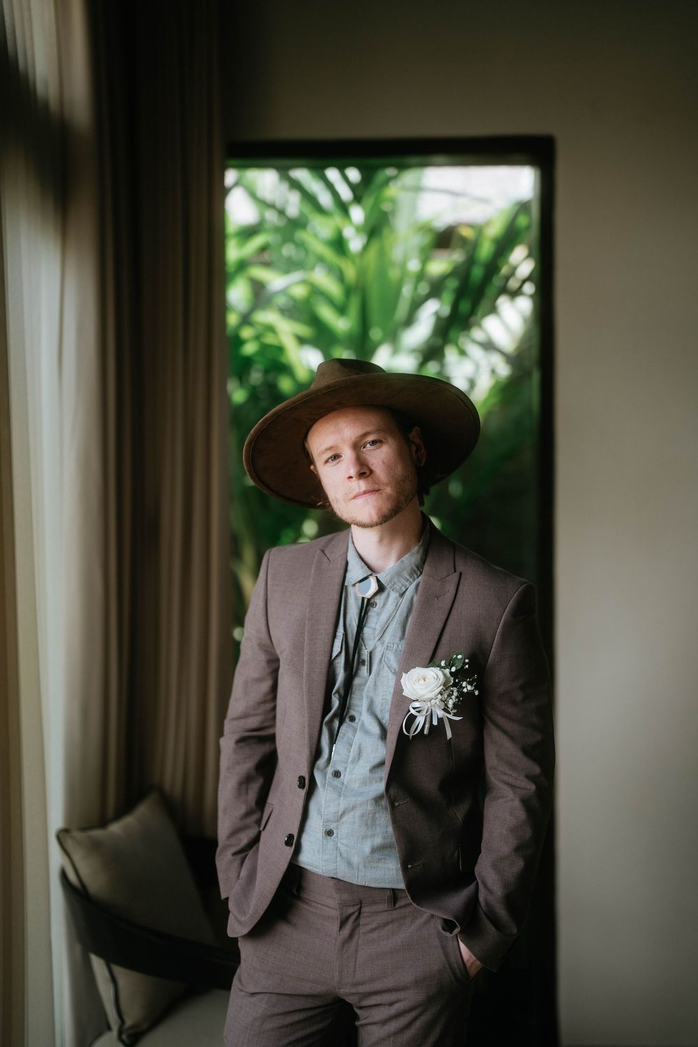 A person in formal attire and hat poses near a window in a tropical setting.