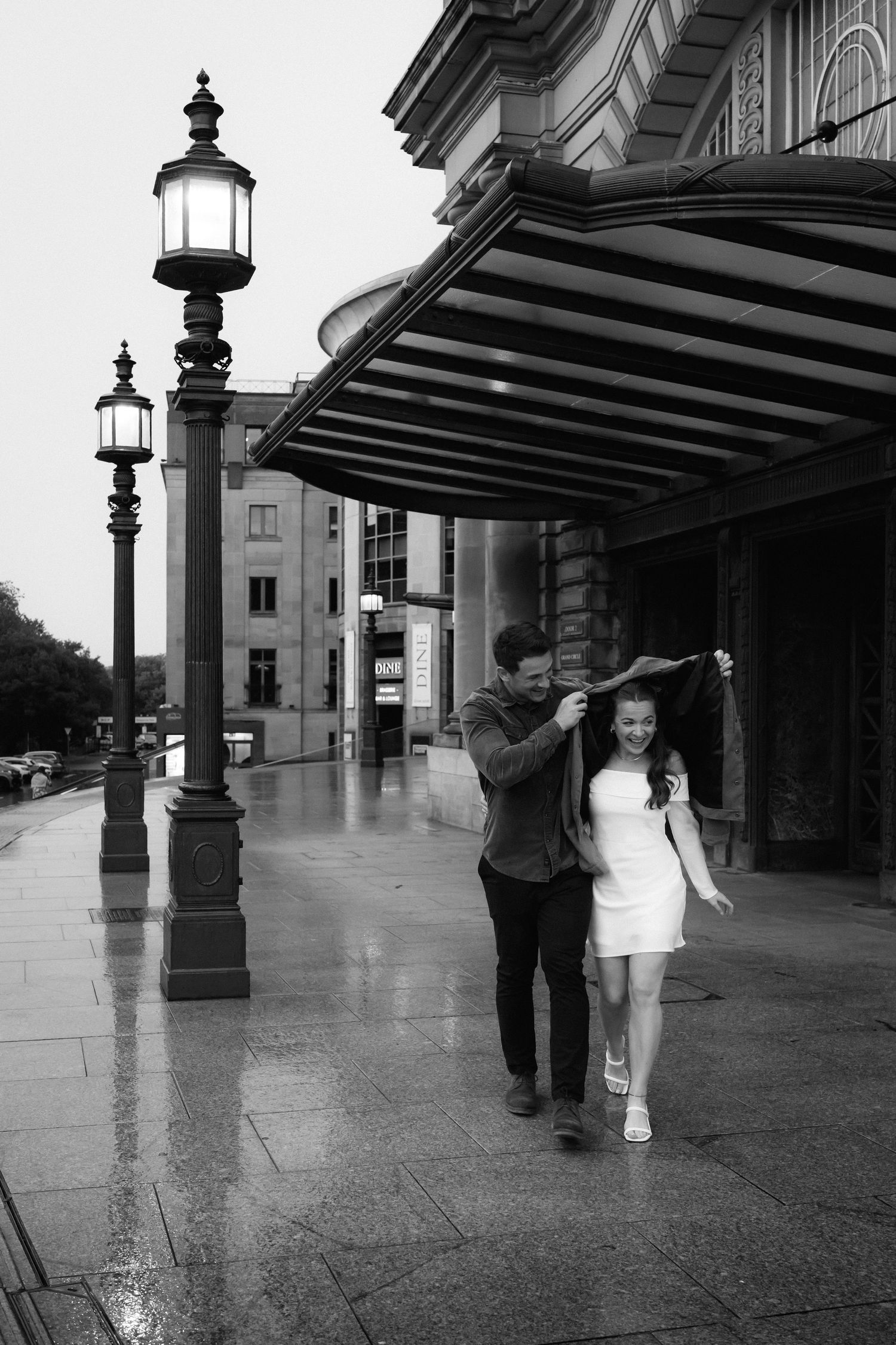 Pedestrians walk through a covered arcade with ornate lamp posts and architecture on a rainy day in black and white.