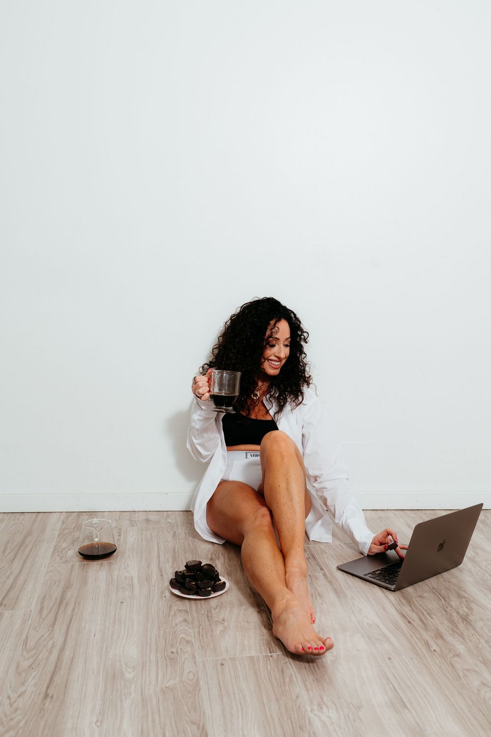 Person in casual attire works on laptop while relaxing on wooden floor with snacks.