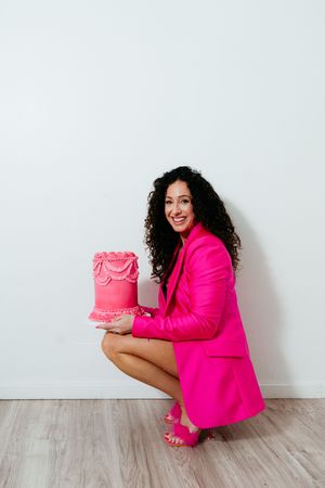 A person in a hot pink blazer poses with a bright pink decorated cake against a white wall.