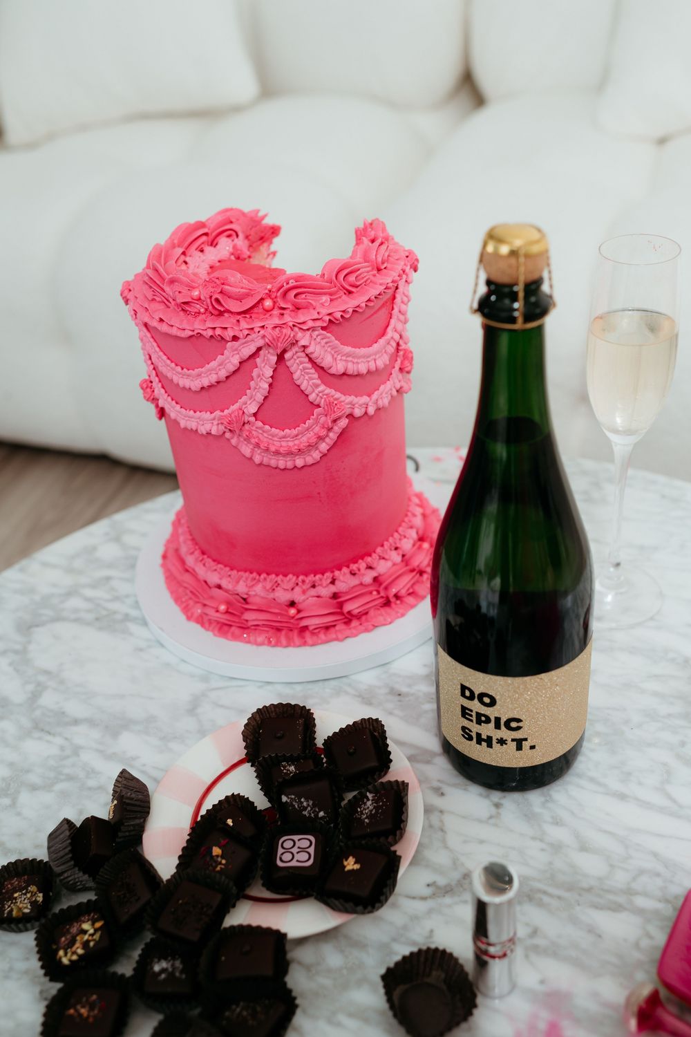 An elegant pink frosted cake with scalloped details sits beside dark truffles and champagne.