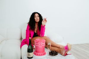 A celebratory scene with a pink birthday cake, champagne bottle, and bright pink blazer on a white couch.