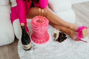 Pink birthday cake with lit candles and champagne bottle on marble coffee table.