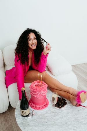 Celebration setup with pink birthday cake, champagne, and matching pink outfit on white couch.
