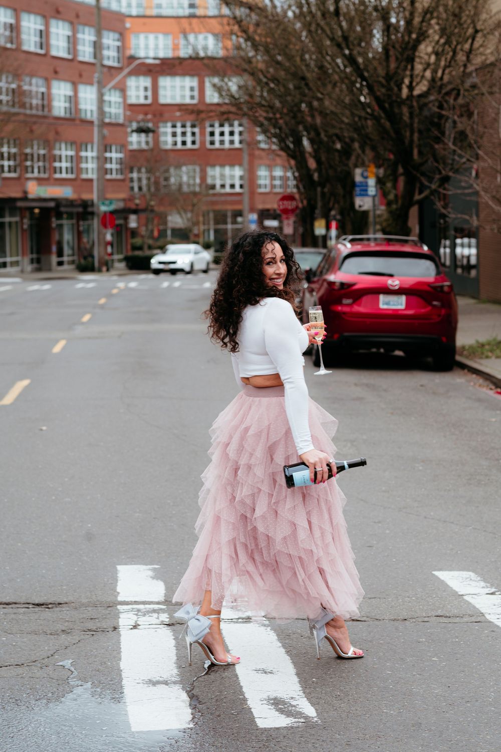 Someone wearing a pink tulle skirt and white top walks across a crosswalk near a brick building.