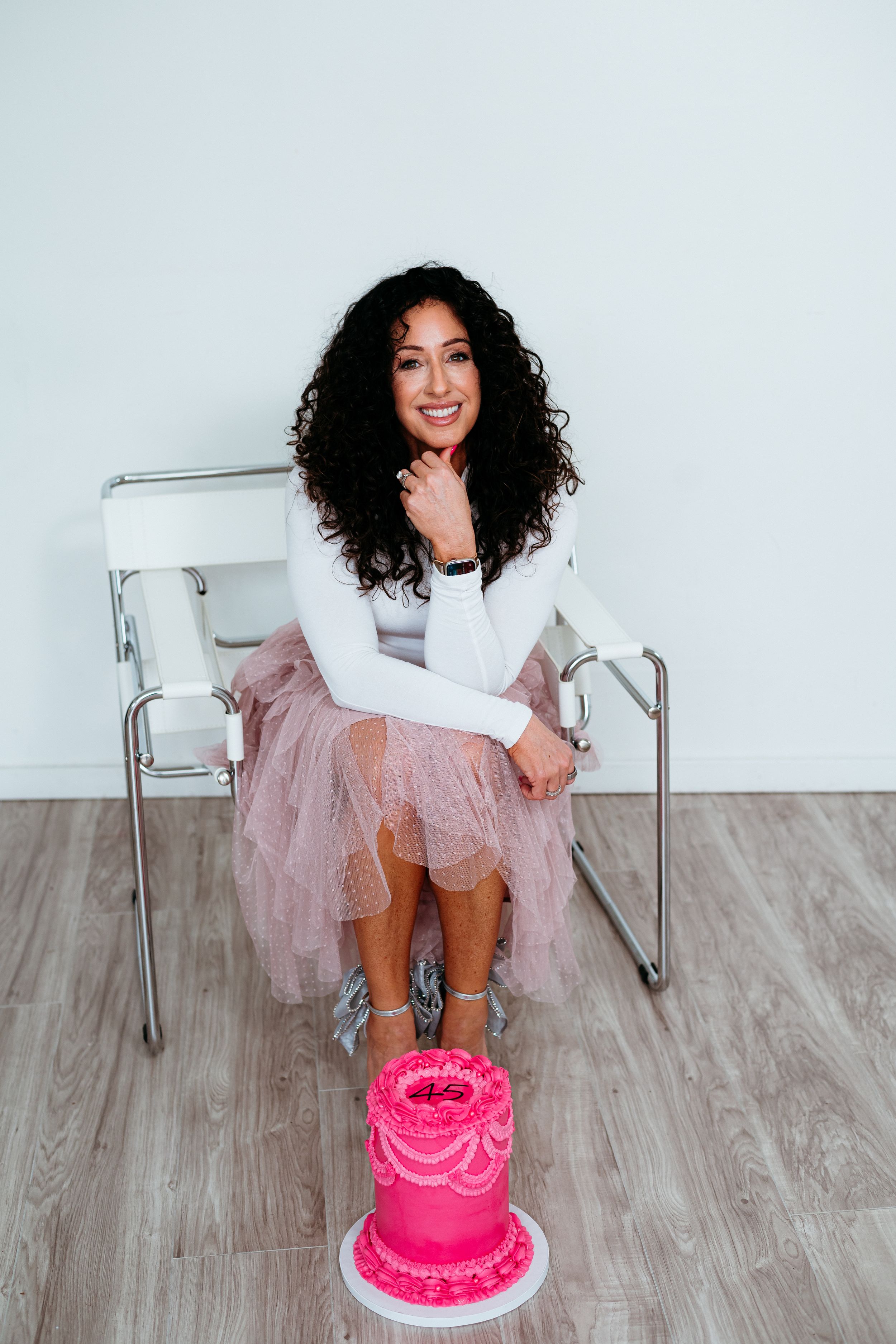 Person in white dress and tulle skirt poses with pink birthday cake on wooden floor.