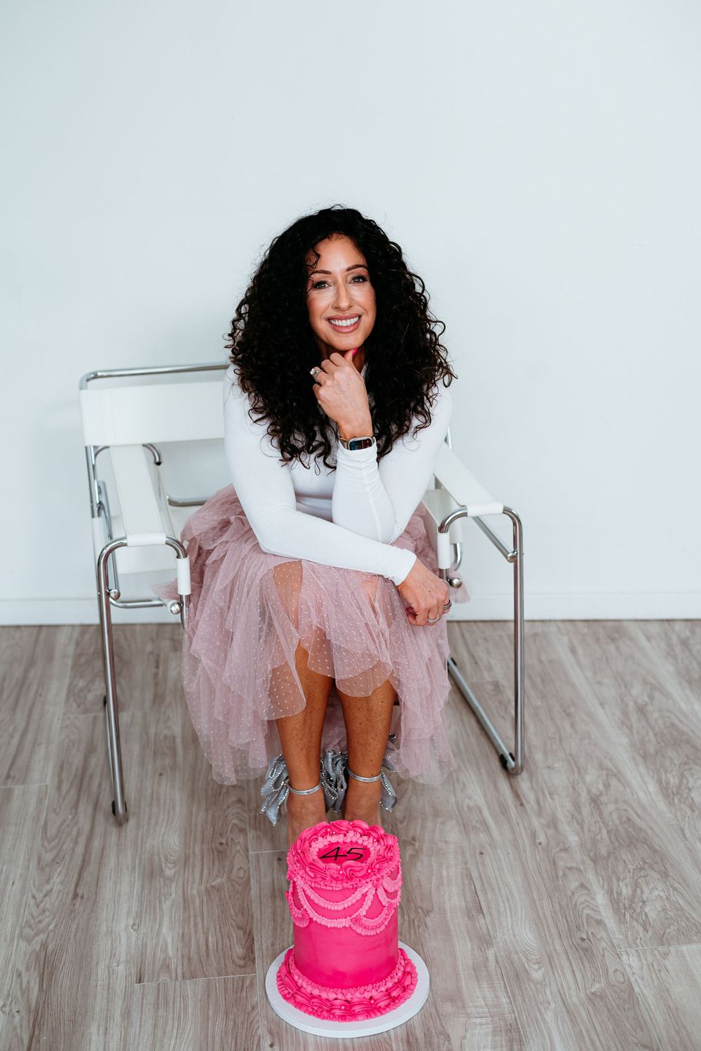 Person in white dress and tulle skirt poses with pink birthday cake on wooden floor.