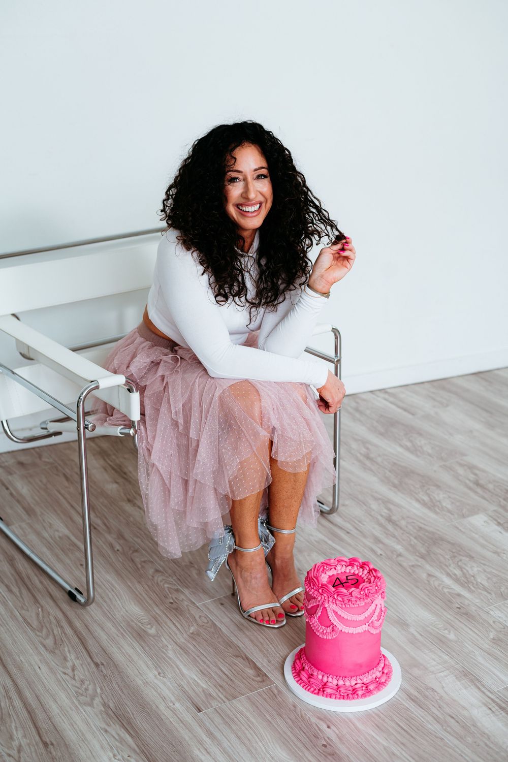 A person in a pink tulle skirt and white top poses playfully near a bright pink birthday cake on a wooden floor.