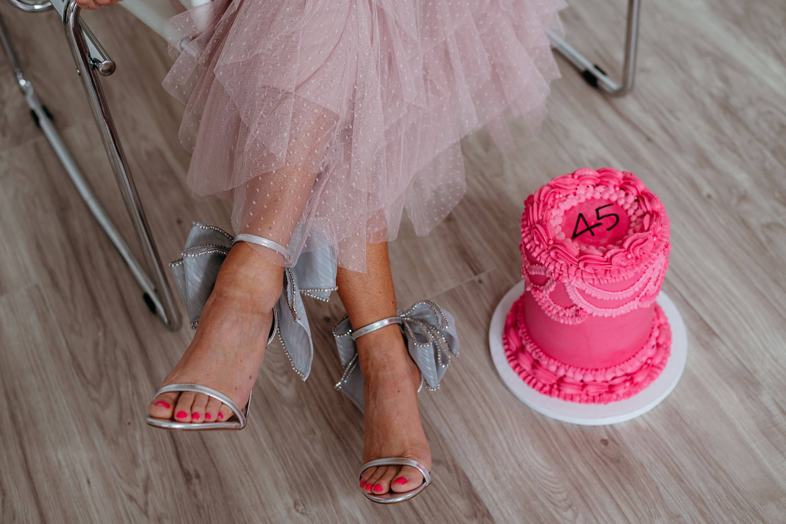 A person in a pink tulle skirt sits on a modern chair next to a vibrant pink frosted cake.