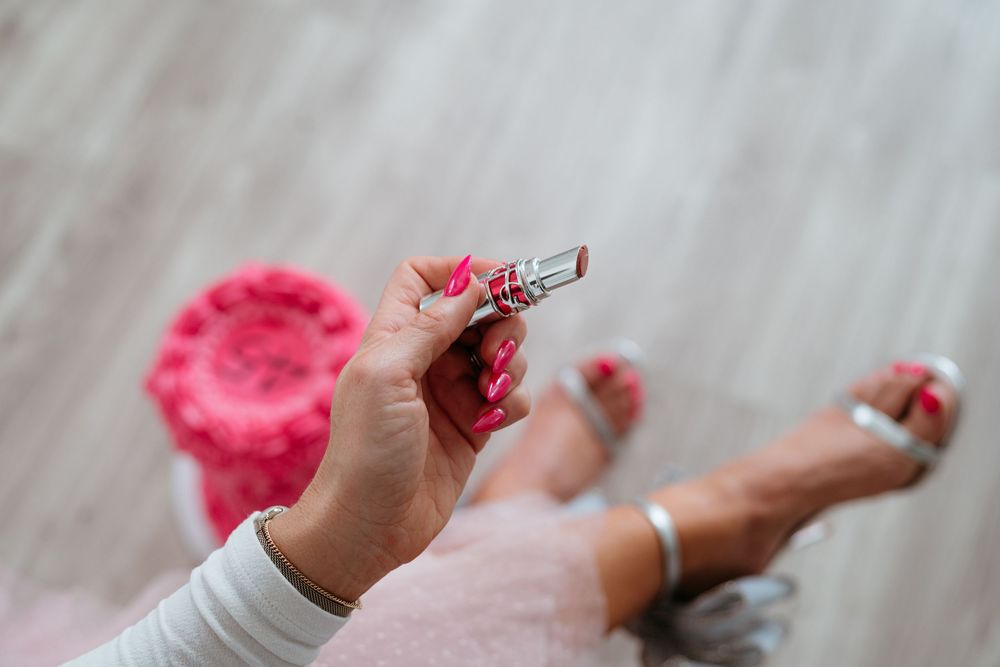 Close-up view of feet in silver sandals next to a pink birthday cake on light wood flooring.
