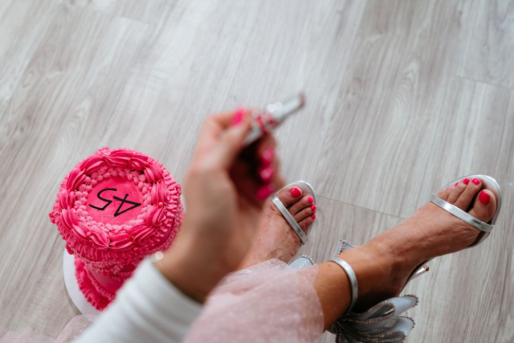 Feet in metallic sandals positioned near a bright pink frosted cake on wooden flooring.