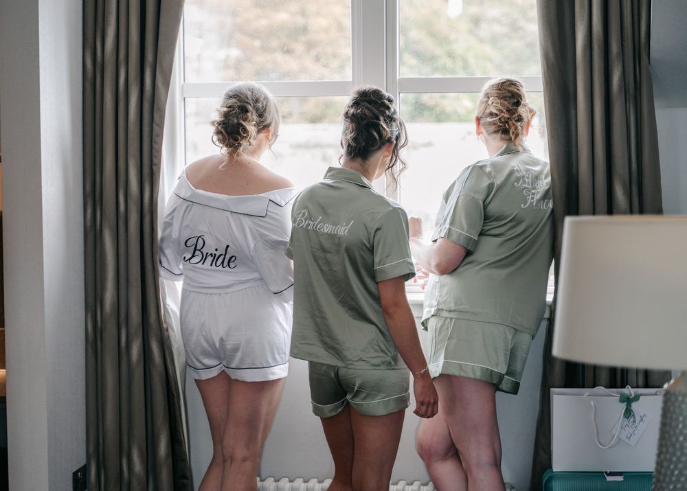 Wedding party members in matching white and sage green outfits stand together by a window.