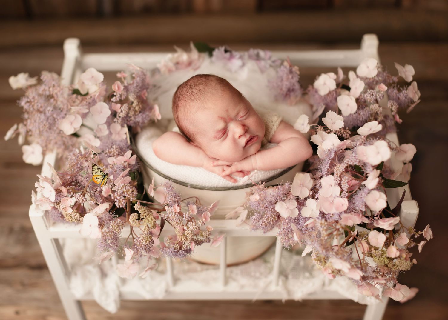 A sleeping newborn nestled among soft pink cherry blossoms and white fabric in a rustic wooden setting.