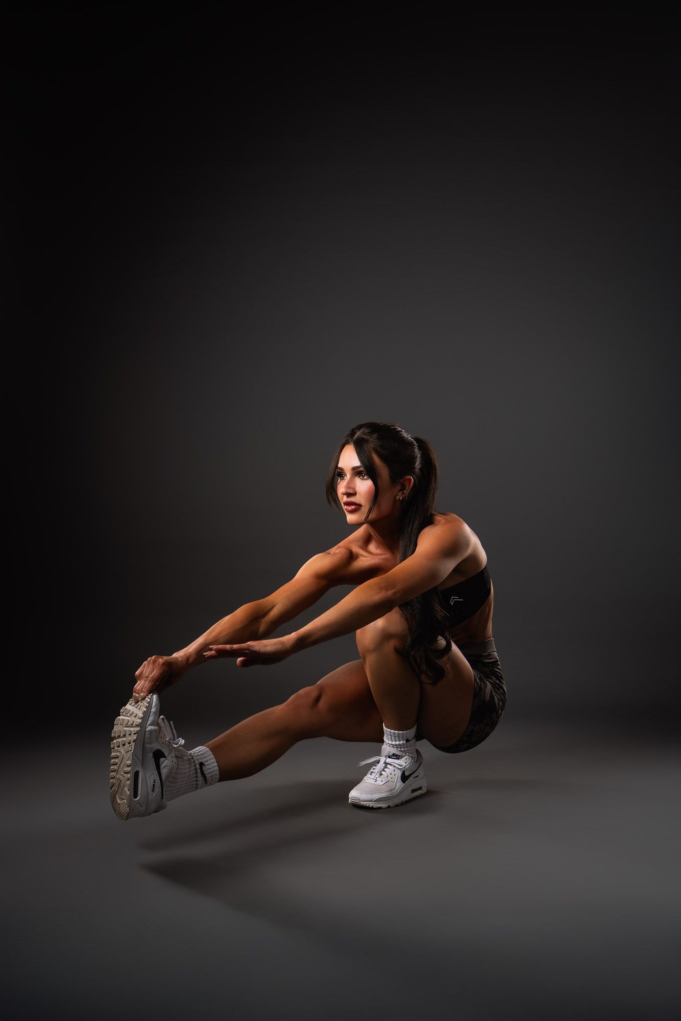 Athletic figure performs dynamic stretching exercises in a dark studio setting wearing white sneakers and sportswear.
