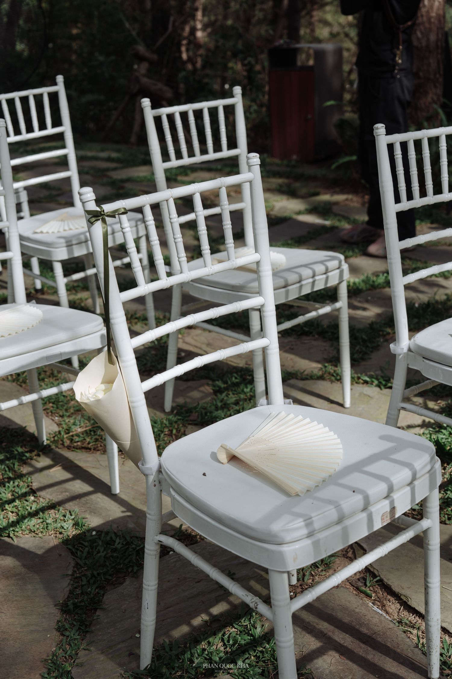 White chiavari chairs with paper fans placed on seats at an outdoor garden wedding ceremony.