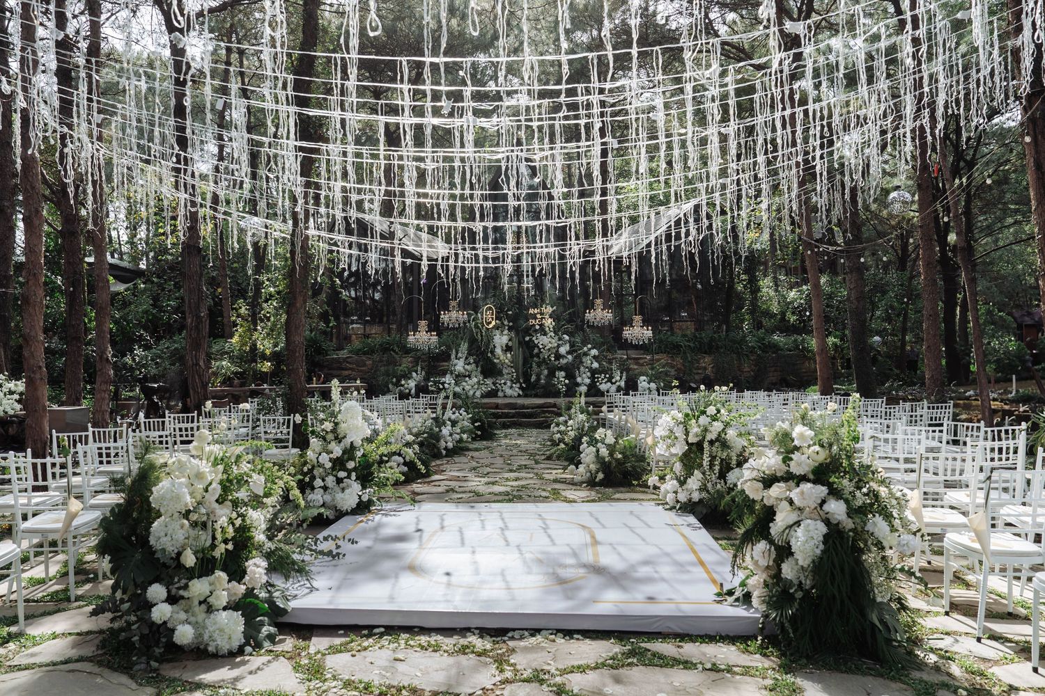 Outdoor forest wedding ceremony aisle with white floral arch, chiavari chairs, and hanging ribbon decor.