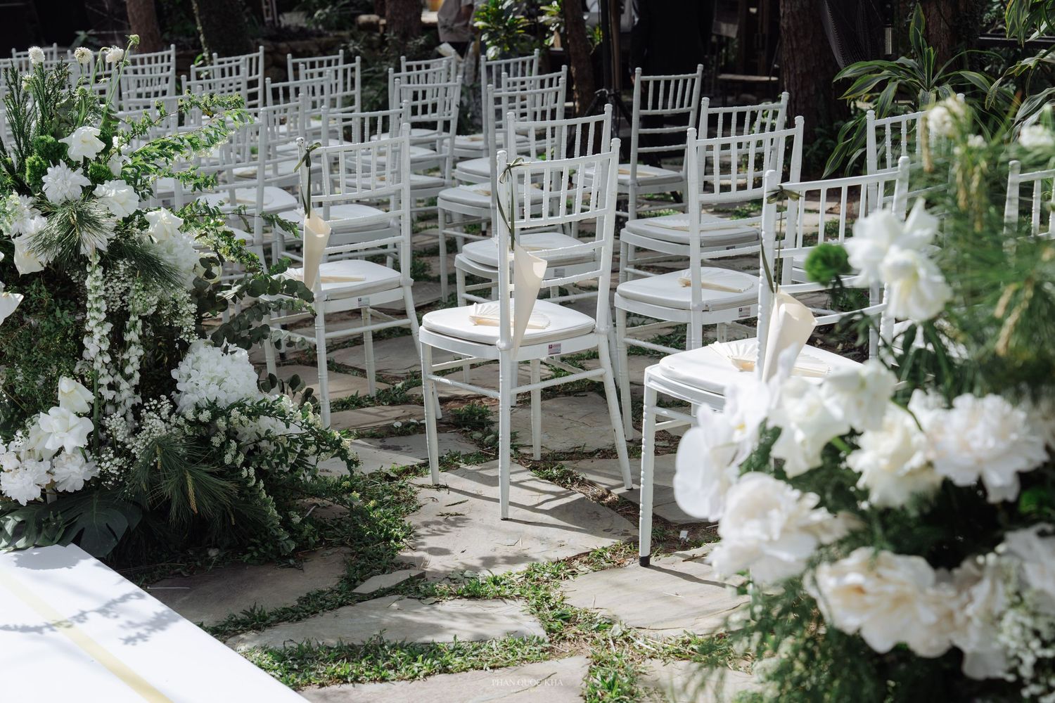 Outdoor wedding ceremony setup with white chiavari chairs lined along a stone path with white flowers.
