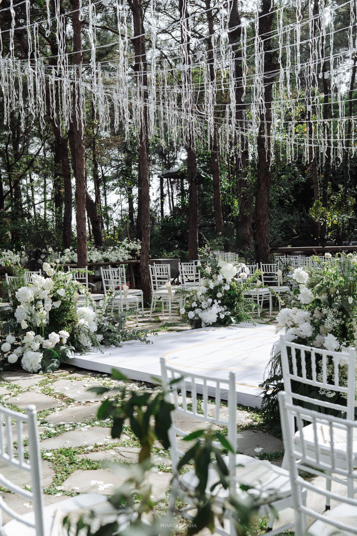 Elegant outdoor forest wedding ceremony with white chiavari chairs, lush florals, and hanging ribbon canopy.