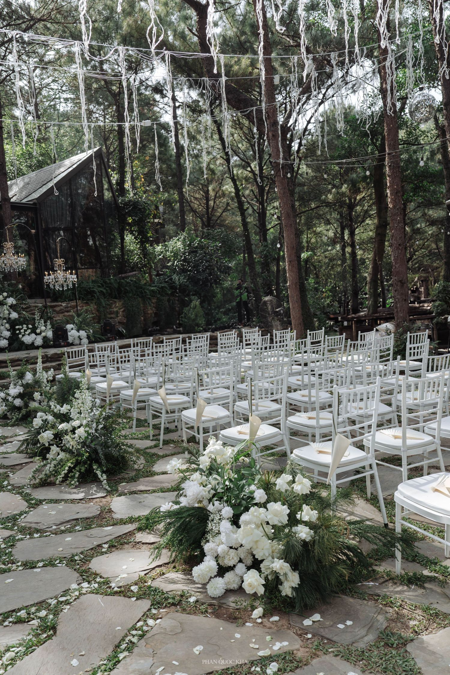 Outdoor forest wedding ceremony with white chiavari chairs, stone path aisle, and white floral arrangements.