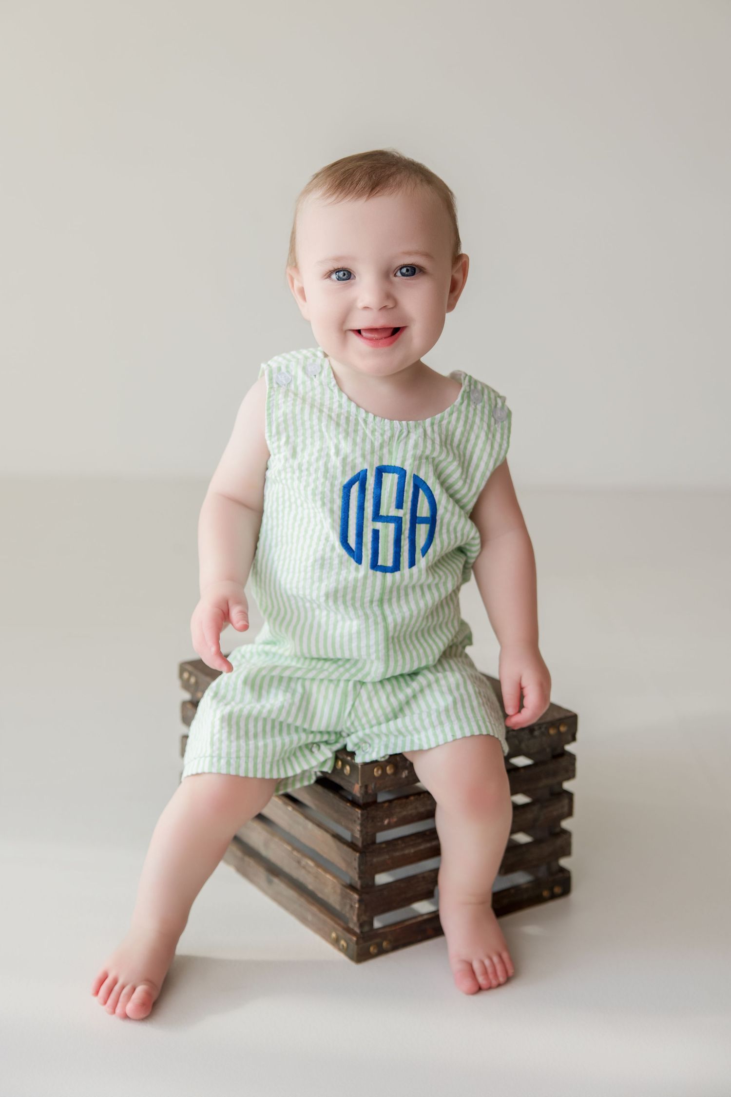 Baby in green seersucker monogrammed romper sits on wooden crate for milestone photo session.