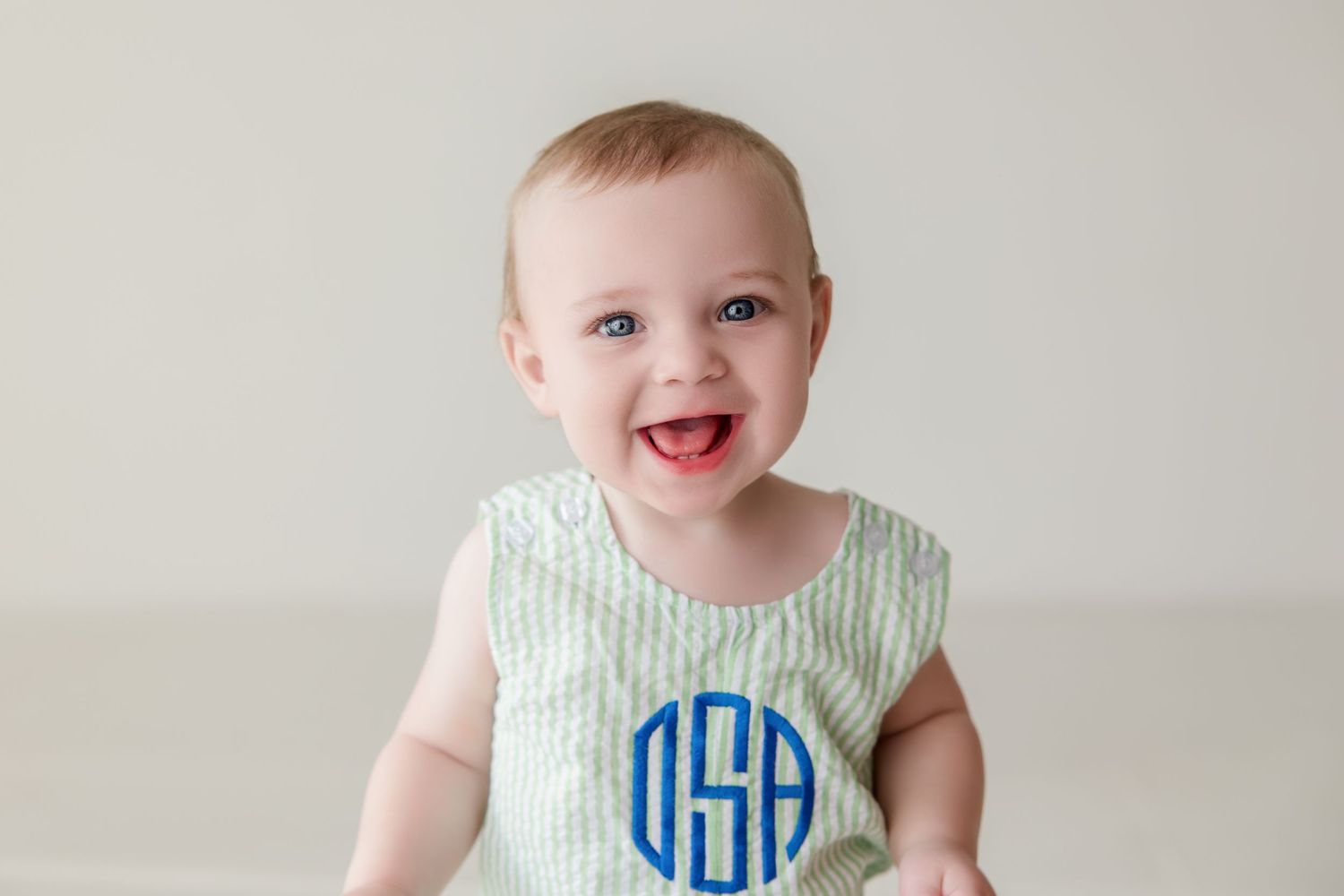 Happy baby wearing mint green monogram romper smiles while sitting against white background.