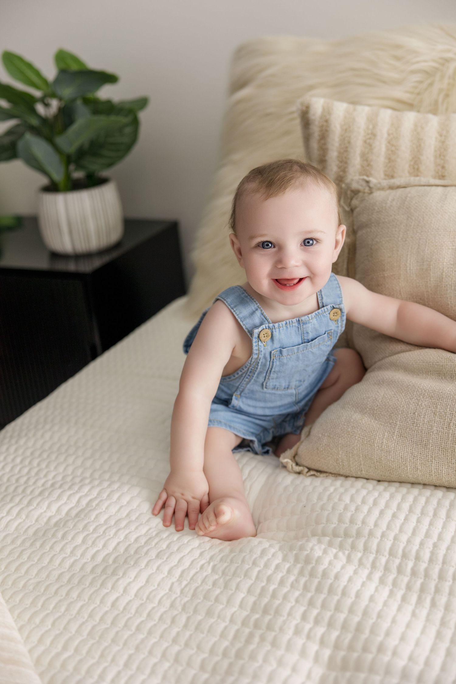 Baby in denim overalls crawls on textured white bedding with decorative pillows.