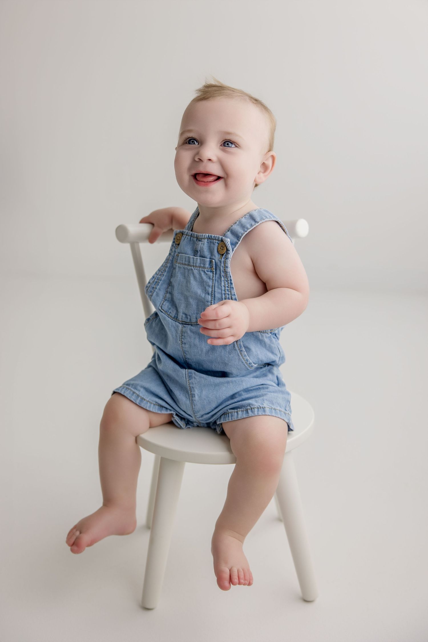 Small child sits on white wooden chair wearing blue denim overalls during photo session.
