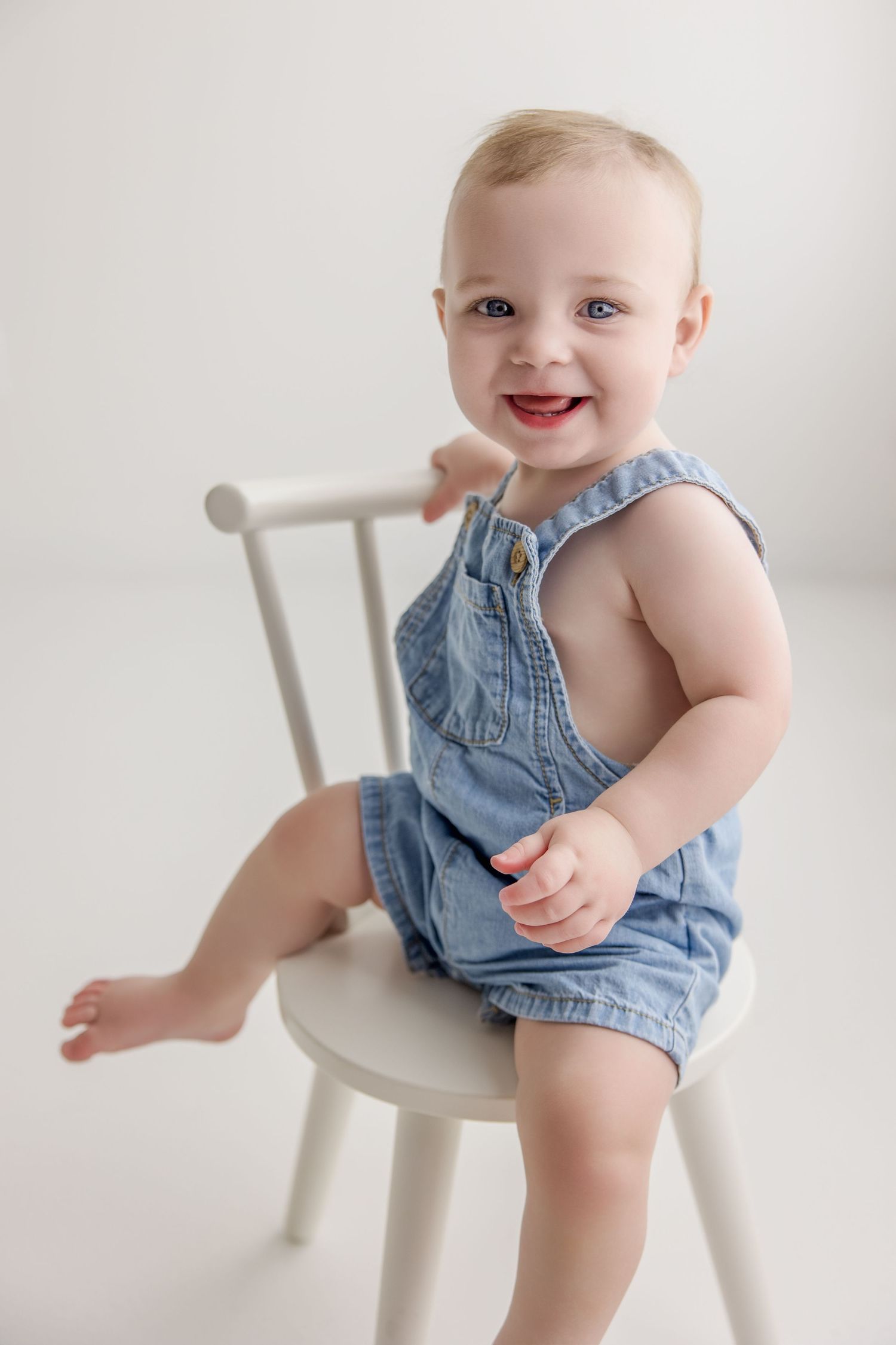 A baby wearing denim overalls sits and plays on a white wooden chair in a bright studio setting.
