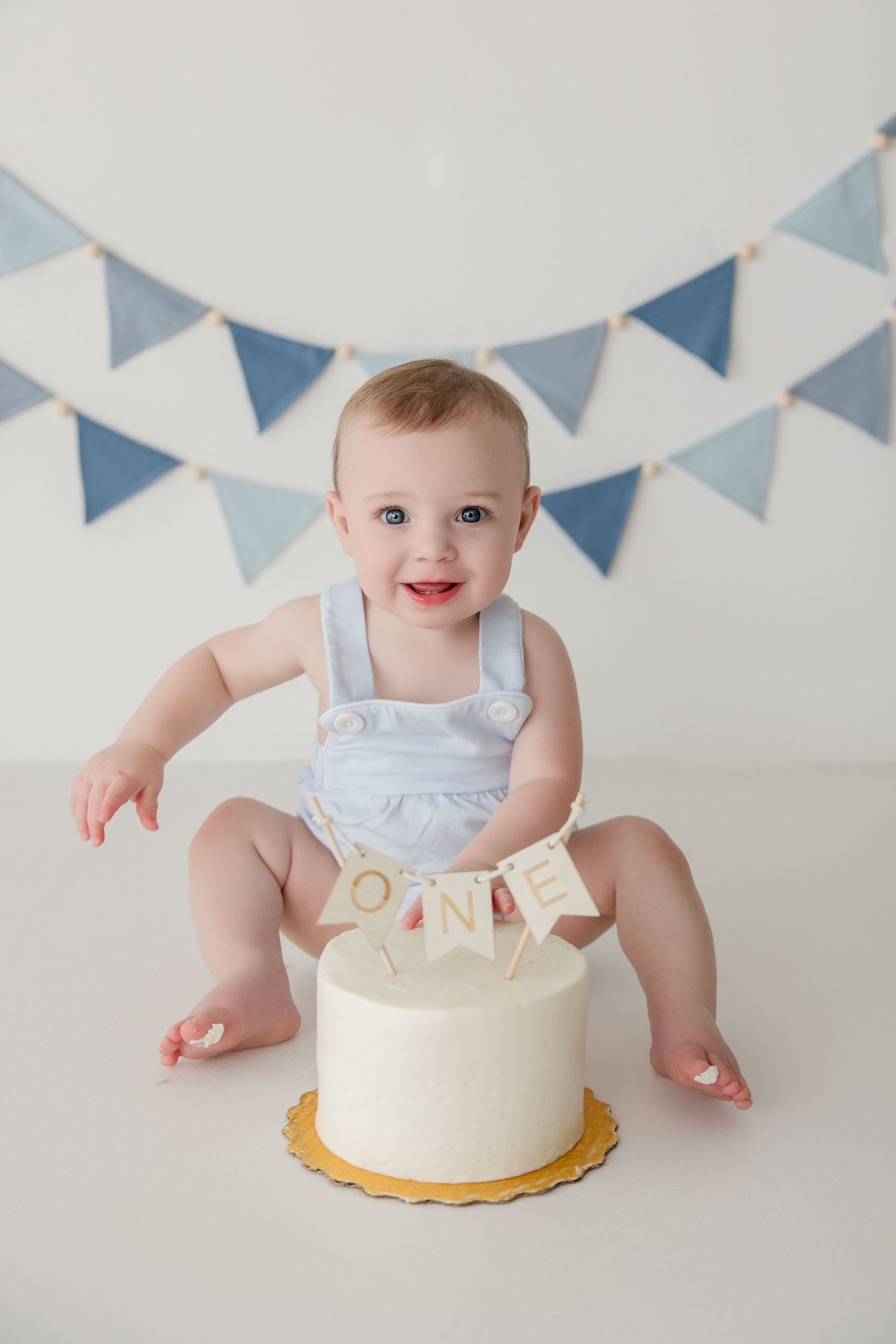 Baby dressed in white sits near a first birthday cake with blue bunting decorations overhead.