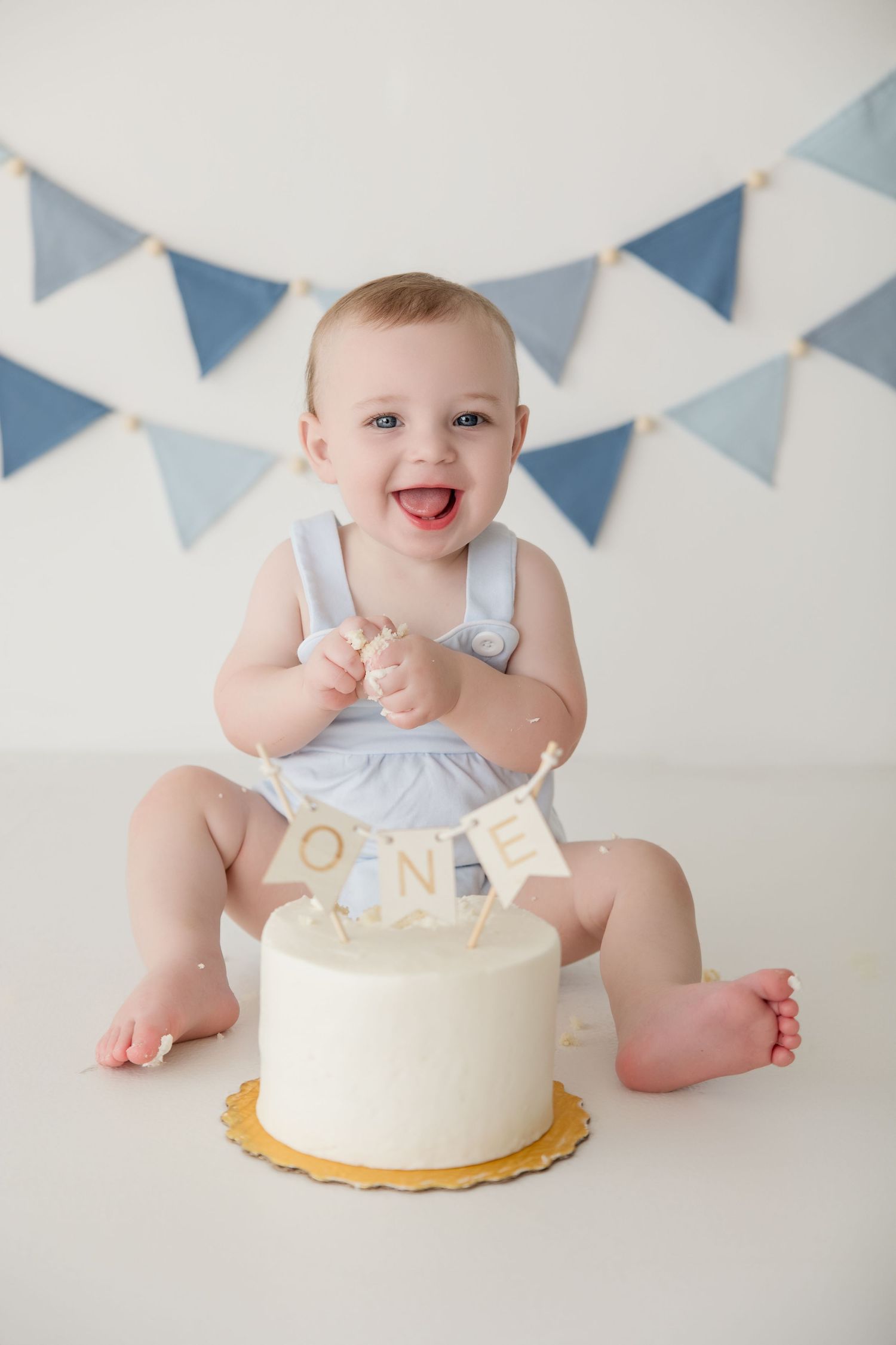 A cake smash photo session featuring a white frosted cake with blue bunting decorations hanging above.