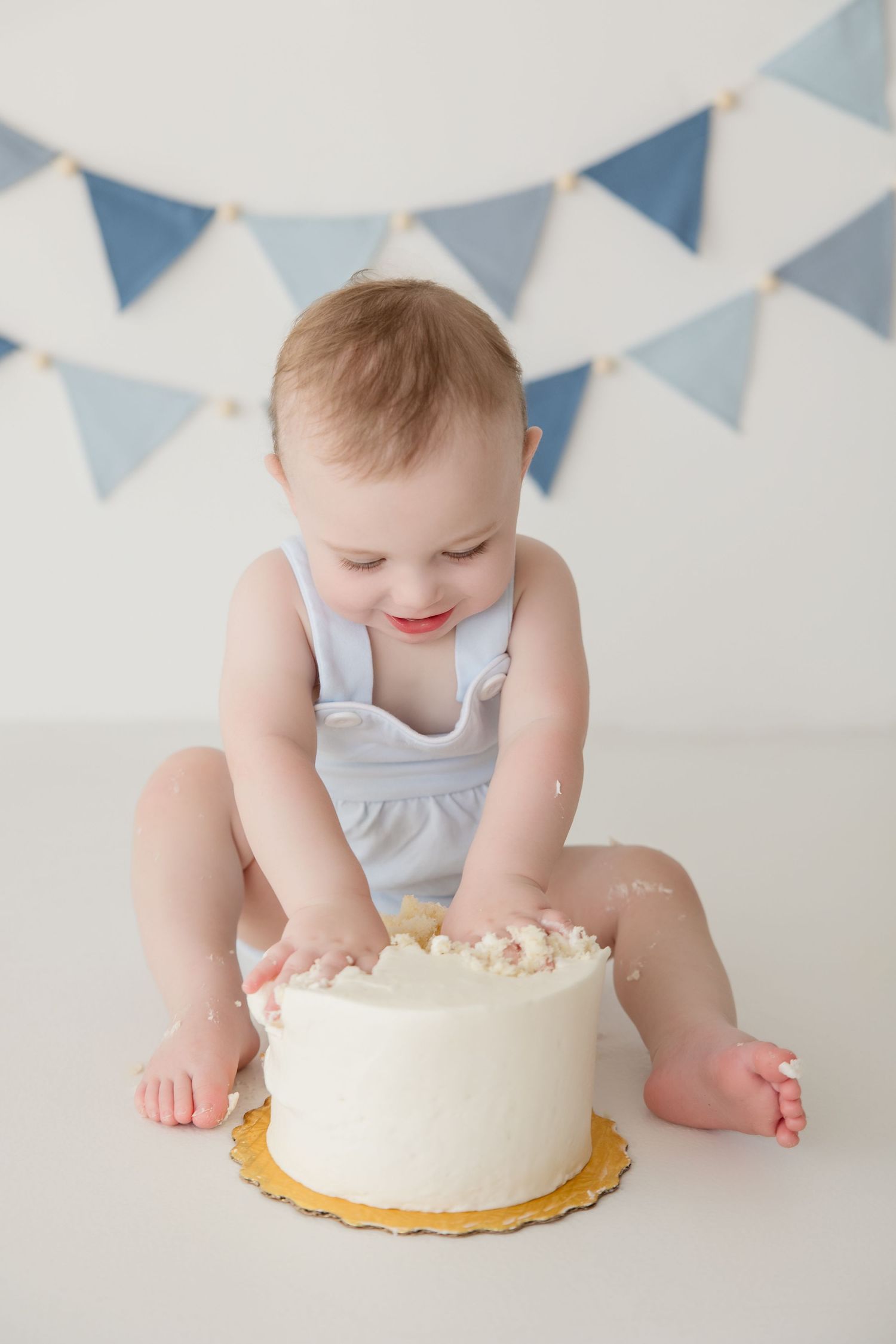 Baby in white outfit reaches for white birthday cake under blue and white banner decorations.